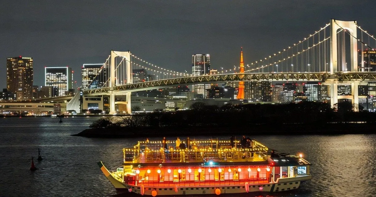 A lit yakatabune houseboat on Tokyo Bay at night in front of the illuminated Rainbow Bridge and the Odaiba skyline